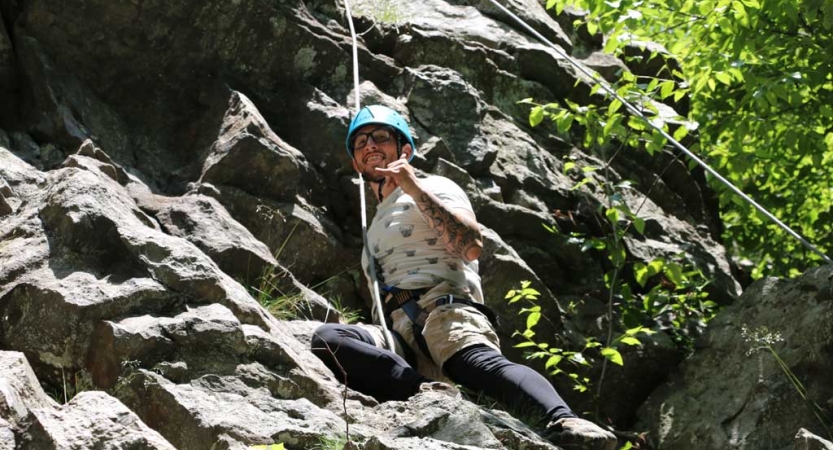 A person wearing safety gear and secured by ropes pauses a rock climbing exercise to give the camera a "hang loose" hand gesture. 
